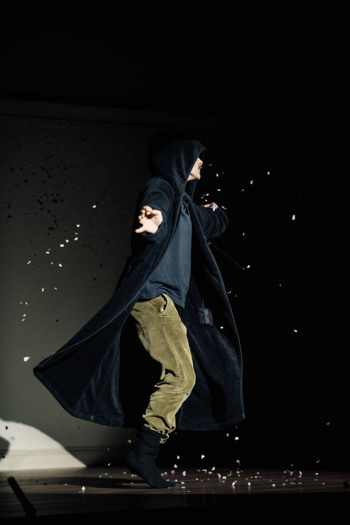 performer spins in a long black hooded jacket on a dark stage